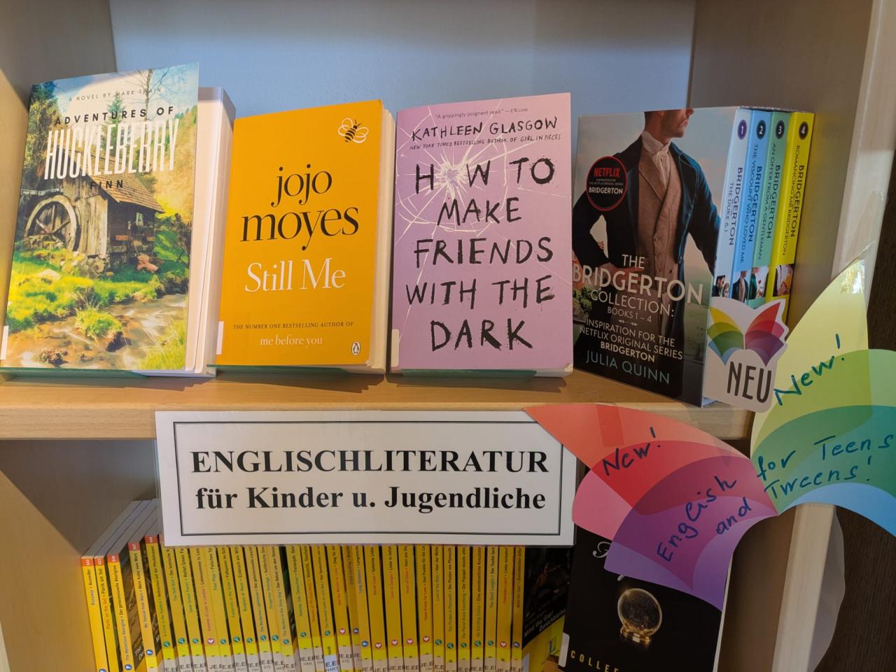 Regal mit neuen englischen Büchern für Kinder und Jugendliche, darunter „Huckleberry Finn“, „Still Me“ und die „Bridgerton“-Reihe, mit Schild „Englischliteratur für Kinder und Jugendliche“ und bunten „New!“-Hinweisen.