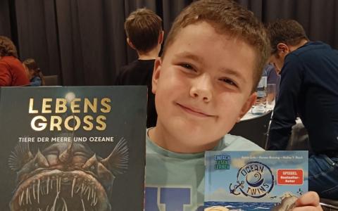 Junge sitzt an einem Tisch und hält stolz zwei Bücher in die Kamera – „Lebensgroß – Tiere der Meere und Ozeane“ und „Dieb-Alarm!“.